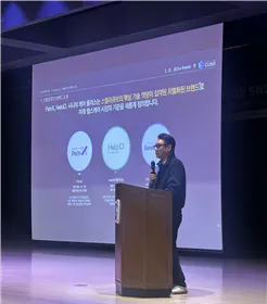 A speaker at a lectern on stage, with a presentation slide behind them showing the three brands PainX, HelciO and Senior Care+